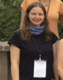 A photo of Lisa Lehmann, a white woman with long brown hair. She wears a black tshirt and a blue bandana around her neck. She also wears a lanyard and name badge from the conference she is attending.